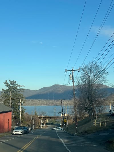 The image presents a forensic, matter-of-fact view of a street descending towards a body of water with mountainous terrain in the background, located in the Town of New Windsor, United States of America. The sky is clear blue with minimal, faint cloud wisps, occupying the upper portion of the frame. Bright sunlight from the left casts long shadows, indicative of either morning or late afternoon.
In the foreground, an asphalt road with a double yellow center line and a white right-side edge line slopes downward and gently curves right. On the left side of the road, a white SUV is parked facing away from the viewer, followed by a dark sedan and a partially visible third dark vehicle. A portion of a red-painted building is visible on the far left, next to a coniferous tree. A utility pole with multiple cross arms and wires stands on the left. On the right side of the road, a white sports car is parked diagonally, facing uphill, near a white crosswalk marking. A prominent wooden utility pole, equipped with a transformer and numerous overhead electrical and communication lines, stands on the right, casting a diagonal shadow. Bare deciduous trees and a grassy slope with scattered vegetation and a low wooden fence are visible on the right.
The mid-ground features a dark metal truss bridge, likely a railroad overpass, spanning above the road. Two yellow circular railroad crossing signs are affixed near the bridge structure. Beyond the bridge, a wide body of calm water, consistent with the Hudson River, stretches across the scene, bordered by a treed and partially barren shoreline on the opposite side.
In the background, a range of large, dark mountains rise, sparsely covered with what appear to be leafless trees. Several antenna towers or communication structures are discernible on the summits of the most distant mountains. No people are visible within the scene.