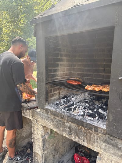 Two men are preparing a barbecue in an outdoor brick grill. The man on the left is wearing a black t-shirt and shorts, and has tattoos on his left arm. He is holding a piece of raw meat. The man on the right is wearing a black baseball cap and sunglasses. He is shirtless and appears to be looking at the meat. Inside the grill, there are several pieces of raw meat cooking on a grate over hot coals. The grill is set into a stone structure. The background shows trees and foliage, suggesting a sunny outdoor setting. The image appears to be taken in Campezo / Kanpezu, Spain.