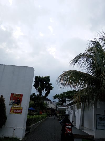 The image depicts a paved pathway in a public area near Alun-Alun Cianjur, Indonesia, under an overcast sky.
On the left side of the pathway, a tall, white, rectangular wall or building segment is visible. Two posters are affixed to this structure: the upper poster is red with white text "WASPADA CURANMOR" (Beware of Motorcycle Theft) and an illustration of a masked figure on a motorcycle; the lower poster is yellow and red, featuring a padlock icon and text "PERHATIAN HARAP JAGA HARTA BENDA ANDA" (Attention, please protect your belongings). Green foliage, including bushes and a small tree, lines the left edge of the path. Further down on the left, a blue tent structure is partially visible.
In the foreground, on the right side of the pathway, a person wearing a dark helmet and dark clothing is seated on a red scooter or motorcycle. The scooter's front wheel and handlebars are visible, and its headlights are off. Further along the pathway, several indistinct figures are visible walking or standing.
To the right, large green palm fronds extend into the upper right quadrant of the image. Beyond these, a white building with modern, angular architectural elements is present. A sign on this building is partially visible, displaying text that includes "PENITIPAN K..." and "ALUN-ALUN CI...", indicating "Penitipan Kendaraan Alun-Alun Cianjur" (Vehicle Storage Alun-Alun Cianjur). Another parked motorcycle is visible further down on the right.
The pathway itself is constructed of dark, interlocking bricks or pavers. The sky is uniformly grey and cloudy, with no direct sunlight visible.
