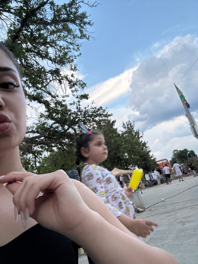 The image captures an outdoor scene in Kostievo, Bulgaria, from a low-angle, close-up perspective. In the immediate foreground, the left side of a woman's face is visible, showing her dark eye, long eyelashes, and slightly puckered lips. Her right hand, featuring long, light-colored nails, rests in the lower-center, appearing to touch her chest. She wears a dark top.
To the right and slightly behind her, a young girl is partially visible, facing towards the right with her head tilted upwards. She has dark hair with a pink clip and wears a light-colored top with a repeating pattern of cartoon characters or emojis. She holds a bright yellow, cylindrical object in her right hand.
The background reveals a lively outdoor setting under a partly cloudy sky with prominent blue patches and large cumulus clouds. Numerous green trees with dense foliage line the middle ground. Further in the background, several casual individuals are present, some walking, others standing near what appear to be market stalls or vendor booths, indicated by light-colored tents and clothing displayed on racks. A tall flagpole with multiple colored flags (including green, white, red, blue, and yellow) stands prominently on the far right. The ground surface is rough, possibly paved, with some scattered debris.