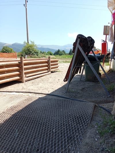 The image depicts an outdoor industrial or construction site, possibly in or near Chelopech, Bulgaria, given the context. In the foreground, a large metal grate lies on the ground, casting a distinct shadow. Behind the grate, a stack of thick, rusty pipes is arranged horizontally, supported by vertical wooden posts. To the right of the pipes, a metal stand holds several large, dark-colored pipe wrenches. Behind the wrenches, a green trash bin and a red fire extinguisher are visible. Further in the background, a telephone pole stands tall against a clear blue sky, with rolling hills and trees in the distance. A safety fence can be seen on the left side of the image, bordering a green field. The ground is covered in gravel and dirt, characteristic of an outdoor worksite. There are no people visible in the frame.