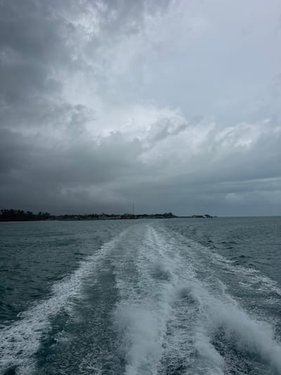 The image depicts a view of the sea, likely taken from a boat moving at speed. In the background is the shoreline of Baan Nai Wok, Thailand, with a faint antenna visible. The sky is overcast with heavy clouds. The turbulent water churned by the boat's wake dominates the foreground.