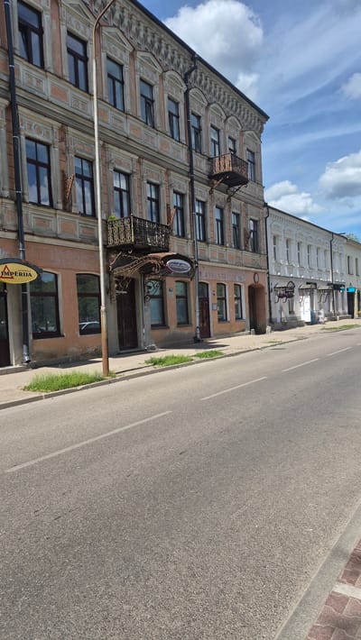 The image depicts a street scene in Daugavpils, Latvia, featuring historic architecture and a paved road. On the left, a multi-story building with ornate detailing and a salmon-pink facade is prominent. This building has dark-framed windows and decorative wrought-iron balconies. A yellow sign with the word "IMPERIJA" is visible on the lower left. Next to it, a light pole stands tall. Further down the street, to the right, another building with a white facade and numerous arched windows is visible. This building appears to be a row of shops or residences. The road in the foreground is asphalt with a white dividing line. The sky above is blue with scattered white clouds. The scene is captured on a bright, sunny day. There are no people clearly visible in the image. The overall impression is of a well-preserved historical urban environment.