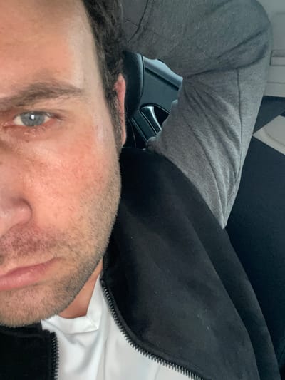 A man is lying down, his head towards the bottom of the frame. He has short dark hair, light skin, and light blue eyes. He is wearing a white shirt with a black jacket over it and grey sweatpants. The background appears to be the interior of a car, with a black seat and part of the steering wheel visible. There is no identifiable location or specific details suggesting this is in the city of Armagh, United Kingdom.