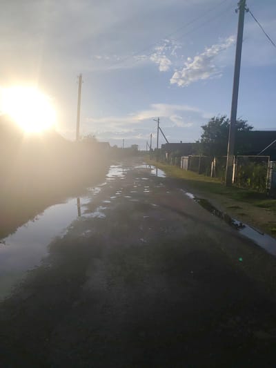 The image depicts a rural road with numerous puddles reflecting the sky and surrounding environment. The sun is prominently visible on the left side, casting a strong glare and creating lens flare. Power lines and utility poles are present, indicating infrastructure in the area. On the right side of the road, there are fences and buildings, suggesting a residential area. The scene appears to be set in the Яглевіцкі сельскі Савет, Belarus, given the context provided. The overall impression is that of a village road after rain, bathed in the warm light of the sun.