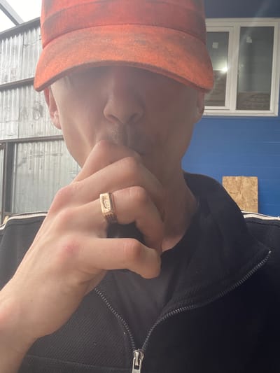A close-up shot shows a person wearing an orange hat and a black jacket. The person's hand is raised to their mouth, and they are wearing a ring on their left index finger. The background appears to be an industrial or outdoor setting, with corrugated metal walls and a blue wall with windows. The image is likely a selfie, given the close-up perspective and the framing of the shot. The specific location in the city of Сокуры, Russia cannot be determined from the provided image.