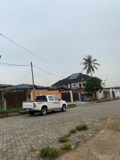 The image depicts a street in Alagba, Nigeria, characterized by paved interlocking concrete blocks. A white Toyota Hilux pickup truck is parked along the right side of the street, adjacent to a row of buildings. One building is tan with a black roof, while another has solar panels on the roof. A palm tree is visible behind one of the buildings. Power lines run along the left side of the street, supported by a wooden pole. Some grass grows between the interlocking concrete blocks. The sky is overcast. There are no people visible in the scene.