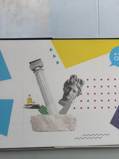 This image shows a poster on a wall next to a gray brick wall. The poster is a collage consisting of a head of a statue in the upper center of the poster and a column protruding horizontally from under the head. The collage also contains a yellow triangle in the upper left, a blue triangle on the bottom, and geometric shapes. A hand is holding a bottle of yellow liquid. The poster features a white grid-like background. The location is likely in Spata-Loutsa Municipal Unit, Greece.