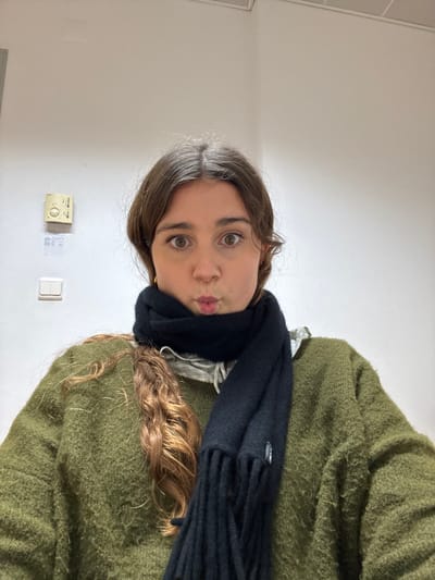 An indoor, waist-up image depicts a young Caucasian female posing in Porto Salvo, Portugal. The subject, with medium brown wavy hair parted centrally and wide brown eyes, looks directly at the camera with her lips pursed. She wears small gold hoop earrings, a thick olive-green knit sweater, and a light-colored ruffled collar shirt visible beneath a tightly wrapped black woolen scarf. The scarf has visible fringed ends and a small light-colored tag. The background is a plain off-white wall. On the left side of the frame, a beige wall-mounted thermostat with a circular dial and two vertically aligned white electrical switches/outlets are visible. Several small, affixed paper labels are present below the thermostat. A white ceiling tile is discernible at the top edge of the frame.
