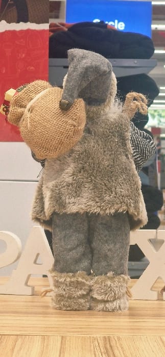 The image, captured in Vendin-le-Vieil, France, presents a rear view of a static decorative figure, resembling a gnome or elf, positioned on a light wooden surface. The figure wears a grey felt conical hat with a small brass bell at its tip, a shaggy light brown faux fur vest, and grey felt trousers. Its lower legs are accented with light brown faux fur cuffs or boots. A burlap sack, containing small, light-colored objects with red ribbons, rests on its left shoulder, and a smaller black and white patterned bag is visible over its right shoulder. To the immediate left of the figure, partial white decorative letters, "P" and "A", are discernible on the wooden surface. The blurred background features a vertical red element on the left and a blue illuminated sign displaying the word "Circle" partially in the upper right, indicative of a retail display environment.