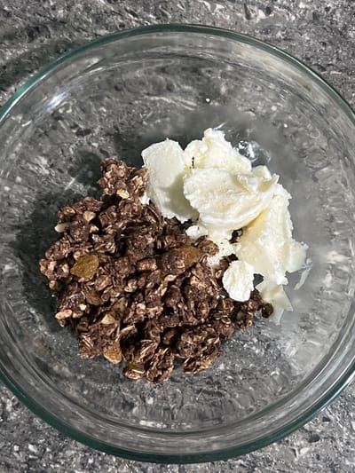 A clear glass bowl filled with chocolate granola and scoops of white yogurt sits on a speckled grey countertop. The granola is dark brown and appears to contain oats, nuts, and possibly raisins. The yogurt is piled to the right of the granola, with some of it spilling over the edges. The scene is shot from a top-down perspective, with the bowl centrally located. The background is out of focus, showing the texture of the countertop. There are no people or animals present in the image. The location in the city of Cuyo, Philippines is not discernible from the image itself.