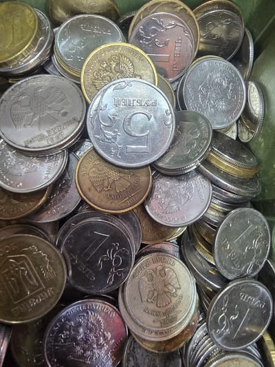 The image is a close-up, overhead view of a pile of Russian coins, predominantly rubles, in varying denominations and conditions. The coins are stacked and scattered within what appears to be a container, with a hint of a green surface visible at the edges, suggesting it is placed in a box or drawer.

The denominations visible include 1 ruble coins, 5 ruble coins, and 10 ruble coins, along with some older or less distinct coins. Some coins display the double-headed eagle emblem of Russia, while others show Cyrillic text and numerical values. The coins are made of different metals, with some appearing silver-toned, others brass-toned, and some showing signs of wear and oxidation.

There is no discernible indication of the city of Nova Kakhovka, Ukraine, or any specific geographical location within it. The image exclusively focuses on the coins. No people, actions, or interactions are depicted. The environment is confined to the pile of coins and the container they are in.