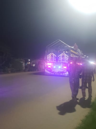 The image depicts a vehicle with illuminated lights in Alalubosa, Nigeria. The vehicle has a rack structure on top. Two individuals are near the vehicle. The scene is at night on a paved surface, possibly a road. Vegetation is visible on the right side of the image.