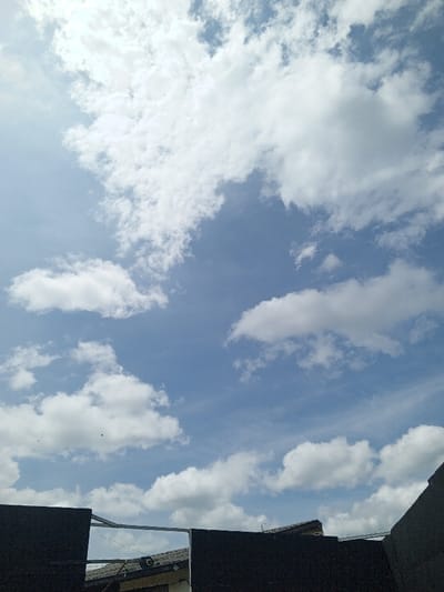 A daytime scene shows a bright blue sky partially obscured by numerous fluffy white clouds. The clouds vary in size and shape, scattered across the expansive sky. In the foreground, a dark, undefined structure with some gray and white details below intrudes from the bottom edge, possibly a building or rooftop. A thin, silvery line runs across the dark structure.