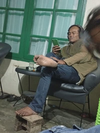 A man sits in a black chair wearing headphones and looking at a green phone. He has one bare foot resting on a small, makeshift wooden stool, and the other leg is crossed at the knee, with his other bare foot visible. A small table with a glass and cigarettes sits next to him. A green window with white curtains is behind him.