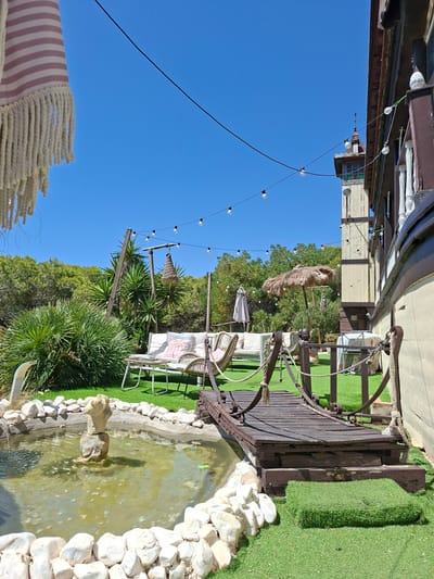 The image shows an outdoor patio area with a fountain, a small wooden bridge, and a seating area, located in La Mata, Spain. The fountain is in the foreground, surrounded by white rocks. A small wooden bridge crosses over a portion of the patio. Beyond the bridge, there is a white sofa and two wicker chairs with cushions. A small table sits between the sofa and chairs. In the background, there is a tall, narrow building with a tower, and lush green trees. String lights with bulbs are strung across the patio, and a thatched umbrella is set up near the seating area. The sky is clear and blue.