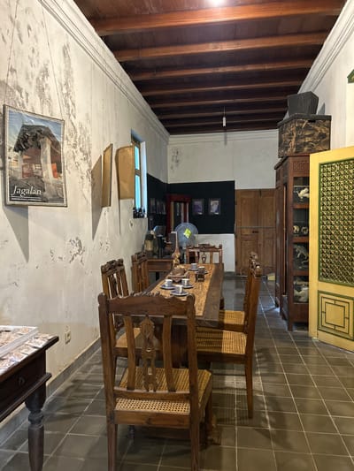 The image depicts an indoor scene, likely a dining area, within what appears to be a heritage building in Banguntapan, Indonesia. The room has a rustic and aged ambiance, characterized by distressed white walls with visible plaster and a dark wooden plank ceiling supported by beams. Sunlight streams in from a window on the left, illuminating the space.

The focal point is a long, dark wooden dining table, set with a placemat, several blue and white ceramic cups, and saucers. The table is surrounded by four wooden chairs with woven cane seats. In the foreground, a dark wooden side table with carved legs holds a display of jewelry or decorative items in a tray.

To the right of the dining area, a tall, dark wooden cabinet stands open, displaying various objects, including what appear to be ceramic or wooden animal figurines and a decorative vase. Next to this cabinet is a tall, yellow door with intricate green geometric detailing. Further back, a wooden door with a horizontal plank closure is visible. The floor is tiled with dark, square tiles laid in a diamond pattern.

The walls are adorned with a framed poster on the left, featuring an image and the text "Jagalan KAMPUNG PUSAKA INTERNASIONAL". Several beige, irregularly shaped objects hang from the ceiling by strings on the left wall. On the back wall, a black expanse is punctuated by three small framed pictures. A vintage-style electric fan is positioned near the center of the room, behind the dining table. The overall impression is that of a well-preserved historical space, potentially a museum or a private residence preserving local heritage.