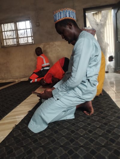 The image shows a man in a blue and white patterned cap and a light blue robe kneeling on a patterned black rug inside a building in Ashaka, Nigeria, at 10.929, 11.475. He is looking down at a cell phone in his hand and his right foot is resting on the floor. Behind him, there are two other men sitting on the rug. The man sitting closest to the camera is wearing a red and orange reflective vest and the other man is wearing a red and orange robe. The image was taken inside a building with a window on the left and a doorway on the right leading to an outdoor space. The interior walls are painted light brown. The lighting is dim, creating a shadowy atmosphere. The image appears to have been taken during the day, but the sun is not visible. The photographer is likely standing to the right of the kneeling man, looking slightly down at him. There is no indication that the man is speaking or interacting with the other men in any way. The mood of the image is contemplative.