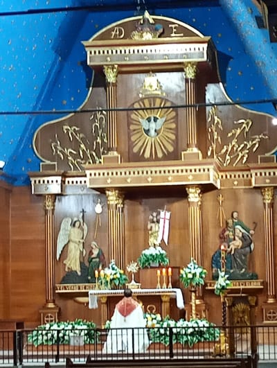 The image captures the altar of the Nuestra Señora del Carmen de La Tirana Sanctuary in Pozo Almonte, Chile. A priest wearing a white and red vestment is kneeling with his back to the viewer in front of the altar. The altar is ornately decorated with religious imagery, including statues of angels, the Holy Family, and symbols of the Holy Spirit. White floral arrangements and candles adorn the altar. The backdrop features wooden carvings and columns with golden accents. The sanctuary's ceiling is painted a deep blue with scattered gold stars. A black metal railing separates the altar from the rest of the space.