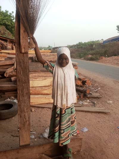 A young girl wearing a white hijab and a patterned green and brown dress smiles while standing next to a wooden structure. She holds a bundle of twigs resembling a broom. Behind her, a stack of lumber and a black tire are visible. The background includes a road, some logs, and distant buildings under a hazy sky. The ground is dirt and small debris.