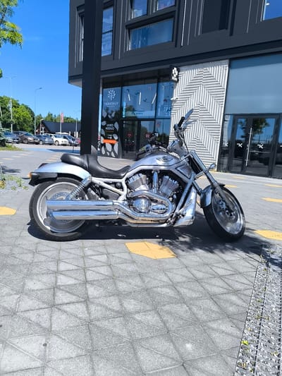 A silver Harley-Davidson V-Rod motorcycle is parked on a paved sidewalk in front of a modern building. The motorcycle is the primary subject, showcasing its chrome accents, large engine, and custom exhaust pipes. It is angled towards the right, with its front wheel turned slightly. The building behind it has a dark facade with large glass windows and a distinctive white, patterned wall section. To the left of the building, a parking lot with several cars is visible, along with trees and a clear blue sky, indicating a daytime scene. The ground is paved with square tiles, some of which are highlighted with yellow markings. The weather appears sunny. Visible text on the building includes "TABASALU ALEVIK", "TABASALU KESKUS", and "UUS KODU", suggesting the location is Tabasalu Center, a shopping or commercial area in Estonia. The overall impression is a bright, clear day with a focus on the parked motorcycle.