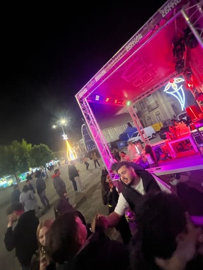 The image shows a night scene in Santo Isidoro e Livração, Portugal. A stage with "CLUB EVENT SPECIAL EDITION" written on it is illuminated with purple and red lights. People are gathered in front of the stage, and streetlights and decorative lights are visible in the background along with some trees. There is a mix of people standing and walking in what appears to be a public space.