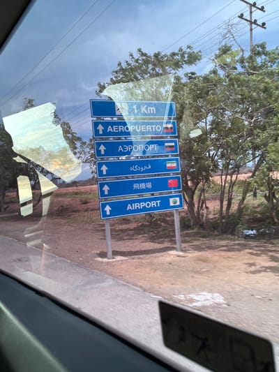 The image depicts a roadside scene in Las Bermúdez, Venezuela, viewed from inside a vehicle through a window exhibiting reflections. The central focus is a blue multi-lingual road sign indicating "Airport" and a distance of "1 Km," mounted on a metal pole on the right side of the road.

The sign consists of six stacked panels, each with an upward-pointing white arrow. From top to bottom, the panels display:
*   "1 Km" (partially obscured by reflections).
*   "AEROPUERTO" (Spanish for Airport), accompanied by a flag that appears to be the Venezuelan flag (yellow, blue, red horizontal stripes).
*   "АЭРОПОРТ" (Russian for Airport), accompanied by the Russian flag (white, blue, red horizontal stripes).
*   "فرودگاه" (Persian/Farsi for Airport), accompanied by the Iranian flag (green, white, red horizontal stripes with the emblem).
*   "飛機場" (Traditional Chinese for Airport), accompanied by the Chinese flag (red field with five yellow stars).
*   "AIRPORT" (English), accompanied by a circular emblem depicting a green landmass on a blue background, resembling a globe or an environmental symbol.

The environment around the sign features dry, reddish-brown earth and sparse, green-leafed trees. A dirt or gravel shoulder is visible in front of the sign, adjacent to a light-colored curb or road edge. In the background, power lines and a utility pole are visible against a light blue, hazy sky. The reflections on the vehicle's window partially obscure the upper left portion of the scene, showing distorted light and possibly a silhouette. The bottom foreground shows the dark interior of the vehicle. The scene is illuminated by daylight.