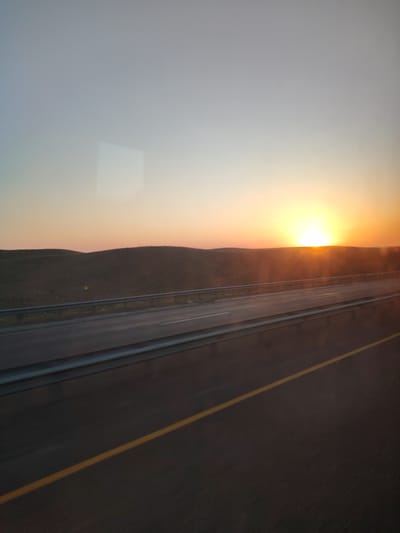 The image depicts a scenic view of a highway at sunset. The sun is low on the horizon, casting a warm, golden glow across the sky and landscape. The sky transitions from a pale blue at the top to a vibrant orange and yellow near the horizon. In the foreground, there is a multi-lane highway, with a yellow line marking one of the lanes. A metal guardrail runs along the side of the highway. Beyond the highway, rolling hills covered in sparse vegetation stretch out into the distance. The overall impression is one of tranquility and vastness, characteristic of a rural or steppe landscape. The image appears to have been taken from inside a moving vehicle, possibly a car or bus, as indicated by the slight motion blur and reflections on what could be a window. The location is stated to be in the city of Қызылсоқ, Kazakhstan, a region known for its steppe landscapes and wide-open spaces.
