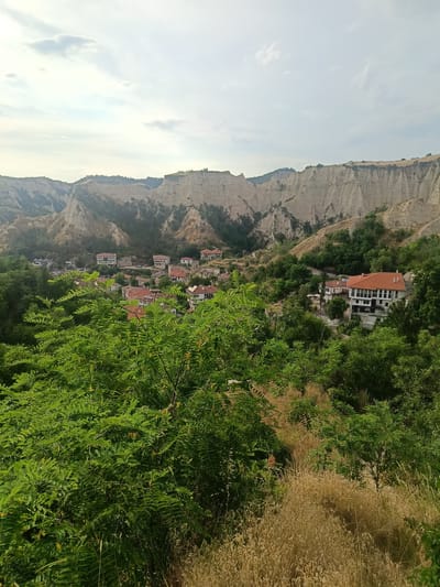 This media file depicts a picturesque view of Melnik, Bulgaria, a town renowned for its unique sand pyramids. The scene is a landscape shot taken from a higher vantage point, looking down into a valley.

In the foreground, lush green foliage, likely trees and shrubs, dominates the lower portion of the image, with some dry, straw-colored grass peeking through. As the view recedes, a charming village with traditional houses comes into focus. The houses are characterized by their terracotta-tiled roofs and white or light-colored walls, many of which feature multiple stories. Several larger buildings, possibly hotels or historical structures, stand out with their distinct architecture.

The mid-ground and background are defined by the iconic Melnik sand pyramids, a series of eroded sandstone formations that create a dramatic and sculpted landscape. These natural geological features rise majestically, displaying their light beige and earthy tones. Scattered trees and greenery are interspersed among the pyramids, softening their rugged appearance.

The sky above is overcast with scattered clouds, suggesting a mild or possibly humid day. The lighting indicates it is likely daytime, possibly late afternoon, given the soft light. There is no visible text in the image. The overall mood conveyed is one of tranquility and natural beauty, highlighting the harmonious blend of human settlement and the unique geological environment of Melnik.