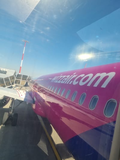 The image depicts a Wizz Air aircraft docked at a jet bridge, likely at an airport. The fuselage of the plane is prominently visible, displaying the "wizzair.com" logo in white lettering against the airline's distinctive pink and blue color scheme. The aircraft's windows are aligned along the fuselage. The sky is bright blue, with the sun causing a glare through the window. A red and white striped pole is also visible, possibly a marker for the jet bridge. The photograph appears to have been taken from inside the aircraft, looking out towards the jet bridge and tarmac. The location is Saint-Louis, France.