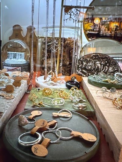 The image shows a display of jewelry, predominantly rings and necklaces, in what appears to be a shop or market stall. The items are arranged on various surfaces, including a rough, textured white stand, a green tiered display, and a dark circular tray. Several pieces of jewelry are tagged with small, brown circular labels, some of which have handwritten numbers or letters. In the background, a snow globe with a miniature building inside is visible, alongside wooden decorative houses and a jar filled with dried organic material. A round mirror reflects another area of the shop, showing more display items and lighting. The overall impression is of a curated collection of artisanal jewelry. There are no people visible in the image. The location "Новинки, Russia" is not discernible from the visual information presented in the image.