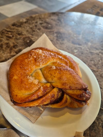 A close-up shot of a cinnamon bun on a small, off-white plate lined with a light brown paper napkin. The bun, glazed and sprinkled with large sugar crystals, is served on a marble-patterned table. A portion of the checkered floor is visible in the background. The bun rests on a table in West Suffolk, United Kingdom.