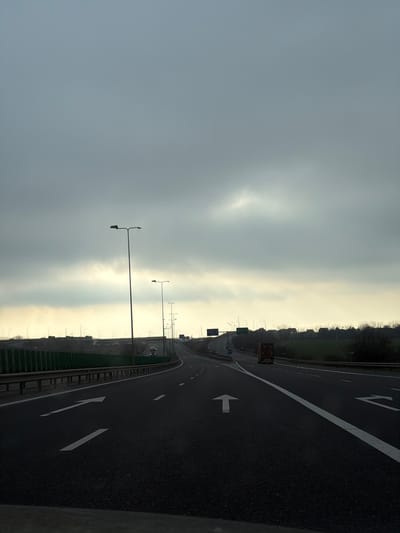 An elevated viewpoint captures a multi-lane asphalt highway traversing the city of Agigea, Romania, under a heavily overcast sky. The sky is predominantly dark grey, with a distinct lighter patch of cloud formation visible towards the upper right. The highway extends from the immediate foreground, where a dark dashboard edge is visible, into the far distance, exhibiting a gentle curve to the right.

The asphalt pavement features clear white lane markings: a solid white line delimits the right edge, dashed white lines separate lanes, and two white arrows are painted on the road surface in the foreground. The left-most visible arrow indicates both a straight-ahead and left-turn option, while the center arrow indicates straight-ahead.

Numerous dark grey streetlights with double-arm fixtures line both sides of the highway, receding into the distance. Along the left side of the highway, a dark metal guardrail is present, beyond which a tall, green, segmented sound barrier runs parallel to the road. Further beyond the barrier, an elevated, undulating terrain with sparse vegetation is visible.

To the right, a similar metal guardrail separates the highway from a flat, green grassy field. In the far right distance, a line of trees or shrubs and scattered low-rise structures are discernible.

Traffic is minimal. One dark-colored passenger vehicle is visible in the left lane in the mid-distance, traveling away from the camera. A large, brown-colored truck with a dark trailer is observed in the right lane, also moving away from the camera. Several overhead gantry signs and pole-mounted road signs are visible in the distance but are illegible. The overall lighting is dim and diffused, consistent with an overcast day.