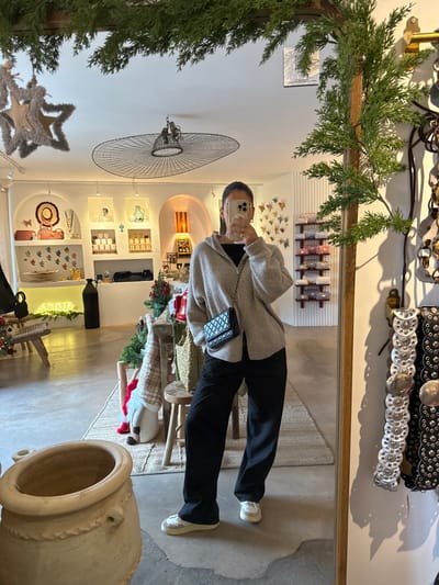 A female subject is depicted in a mirror selfie, taken inside a retail store located in Saint-Paul-de-Vence, France. The subject stands centrally, dressed in a light beige hooded zippered cardigan over a black top, black wide-leg pants, and light-colored sneakers. A small black quilted shoulder bag with a chain strap is worn across her body. Her face is partially obscured by a silver smartphone held up to capture the reflection.

The mirror frame is adorned with artificial green pine branches. To the immediate right, outside the mirror's reflection, hangs a display of dark-colored belts featuring metallic silver studs and discs.

The reflected store interior features white walls with arched display alcoves and shelves showcasing various artisanal goods, including pottery, woven items, candles, and framed art. A large, flat, dark-colored wire-frame light fixture is suspended from the white ceiling. The flooring consists of concrete or polished stone, partially covered by a light-colored woven rug. In the middle ground, a wooden table or counter is decorated with green garlands, red accents, and a small gnome figurine, indicating a festive or holiday theme. A rustic wooden stool stands nearby. Further into the background, a display of rolled textiles is visible on shelves, alongside a wall adorned with numerous colorful butterfly motifs. A tall black cylindrical object and a partially visible neon sign are also present. In the foreground, outside the mirror's reflection to the bottom left, a large, light-colored pottery urn is situated.