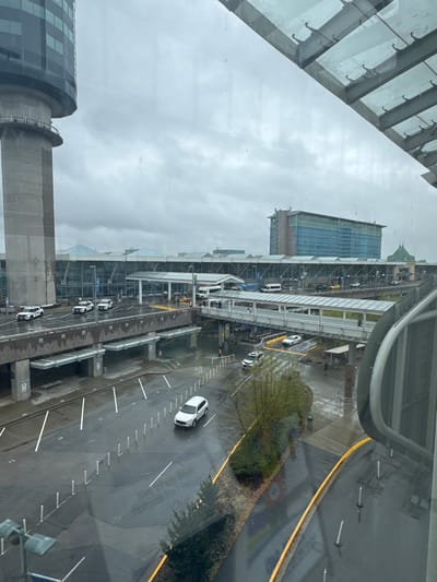 The image is an aerial view of an airport or a similar transportation hub on a cloudy day. The scene is dominated by a large, modern building with a glass facade and a distinctive curved roof. Outside, a multi-level roadway is visible, with several cars driving on it. One white car is prominent in the foreground, and other vehicles, including a white van, are scattered throughout the scene. There are elevated walkways connecting different parts of the complex. The ground is wet, suggesting it has recently rained or is raining, and the sky is overcast. The overall mood is somewhat somber due to the weather. There is some faint text visible on the road surface and on some signage, but it is not legible enough to be clearly read. The architectural style and the presence of airport-like infrastructure suggest a location in a major urban area, consistent with Richmond, Canada, which is known for its international airport.