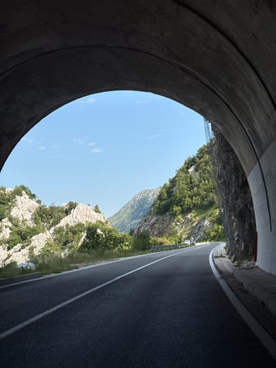 The image captures the exit of a dark tunnel onto a winding mountain road in Dragalj, Montenegro. The tunnel's concrete arch frames a bright, clear blue sky with a few scattered clouds. Outside the tunnel, a paved asphalt road curves to the left, bordered by a metal guardrail on the right side.
The surrounding environment is mountainous and rugged. Steep, rocky slopes are covered in dense green vegetation, primarily trees and shrubs. The terrain is characterized by sharp, white rock formations interspersed with foliage. In the distance, more rounded, tree-covered mountains are visible against the horizon.
There are two white vehicles visible on the road, one further ahead on the curve and another smaller vehicle that appears to be a car closer to the tunnel exit. The road surface is dark asphalt with a solid white line marking the edge of the lane. The perspective suggests the image was taken from within the tunnel, looking out towards the open landscape. The right wall of the tunnel shows a rough rock face overlaid with a protective mesh netting. The interior of the tunnel is dark and textured.