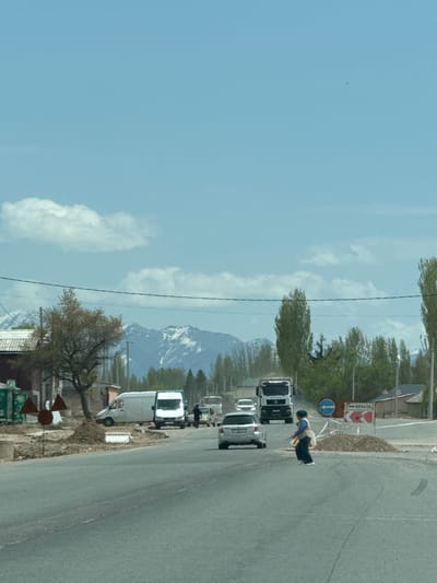 The image captures a daytime scene along a road in a mountainous region, specifically Temir, Kyrgyzstan. The sky is bright blue with scattered white clouds, suggesting clear weather.
In the foreground and mid-ground, a paved road stretches, with several vehicles visible. These include what appear to be multiple trucks and some cars, mostly light-colored. On the side of the road, near the edge of the asphalt, a person is visible. They are wearing dark clothing, a light blue top, and a yellow or orange safety vest or apron, standing near a set of road signs.
Two prominent road signs are present:
1. A red and white rectangular sign with arrows indicating a sharp turn. Above the arrows, the Cyrillic word "ОБЪЕЗД" is written, which translates to "detour" or "bypass."
2. A blue circular sign with a white arrow pointing to the right, typically indicating a mandatory turn or "keep right."
Beyond the road and foreground elements, trees with light green foliage are scattered. In the distance, large mountains dominate the horizon, with several peaks still covered in snow, indicating a significant altitude or early spring/late autumn season. A power line traverses the upper portion of the image.
The scene depicts a typical roadside environment in a mountainous, likely rural or semi-urban, setting.