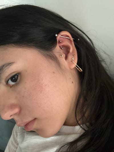 An individual is visible, featuring dark hair and fair skin with visible pores and freckles. The focus is on the ear, adorned with multiple piercings, including a gold safety pin. Various studs and hoops are apparent, one of which bridges across the ear. The subject's eye is open and facing the camera. A white garment appears in the lower-right corner, possibly a shirt or bedsheet.