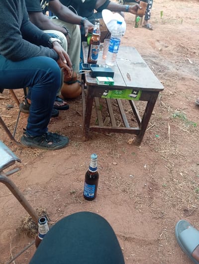 A group gathers outdoors, enjoying drinks around a weathered wooden table. A brown beer bottle and water bottle sit atop the table, along with cigarettes. A calabash gourd rests beside the table. More beer bottles are seen on the ground amongst the red dirt. Multiple people are visible, clad in casual clothing.