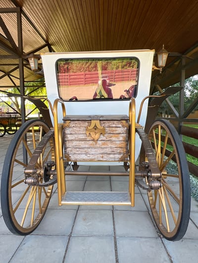 A white horse-drawn carriage with gold accents is parked on a paved surface under a wooden canopy. The carriage features a wooden chest on its rear, and large spoked wheels. The window of the carriage reflects a red wooden fence and greenery. Two gas lamps are mounted on the canopy support beams. The setting appears to be an outdoor venue, possibly a park or historical site, in Plastunivska, Russia. No people are visible in the image.