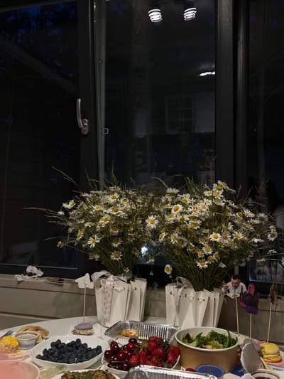 The image depicts an indoor scene, likely a dining area, with a large window in the background reflecting the night sky and trees. On the windowsill, two bouquets of daisies with grass are arranged in white bags tied with pink ribbon. A table in the foreground is laden with food items, including a bowl of blueberries, strawberries, cherries, a salad, what appears to be pastries or bread, and a small dish of sauce. Several decorative elements, including paper cutouts of people, are placed on sticks. The overall atmosphere suggests a celebratory or gathering event. The location is identified as Ivachiv Horishnii, Ukraine.