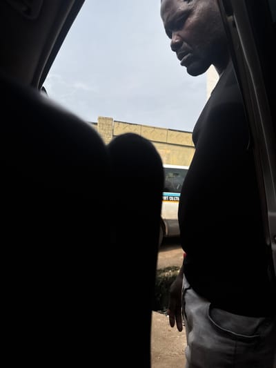 A medium shot captures a Black man in profile, looking downwards. He is wearing a black t-shirt and light grey pants. The man stands near the open door of a vehicle. In the background, a white vehicle with "PORT CO. LTD" visible on its side is parked. The man's left hand rests on his pants pocket. The scene is set outdoors, with a building structure and cloudy sky visible behind him. The image appears to be taken within the city of Orie Uratta, Nigeria.