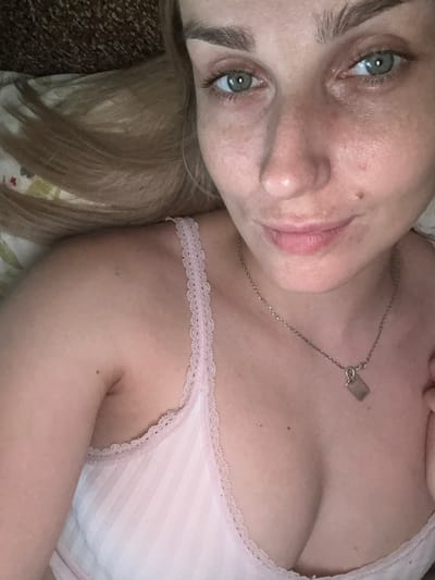 The media file is a close-up, low-angle, portrait-style photograph of a young woman lying down. She has fair skin, light blonde hair, and light blue-green eyes. She is wearing a light pink tank top with white striped detailing and lace trim. A delicate silver necklace with a rectangular tag hangs around her neck. The lighting is soft, suggesting an indoor setting, possibly during the daytime or with artificial lighting. Her expression is relaxed and perhaps slightly playful, with a gentle smile. The background is out of focus, showing hints of fabric with a subtle floral pattern, and appears to be bedding. The overall mood is intimate and casual. No visible text is present in the image. The provided location context of Piatykhatky, Ukraine, does not offer any specific visual cues within the image itself.