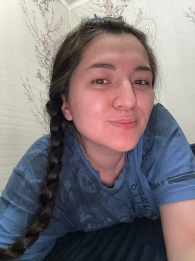 An image displays a single female individual positioned indoors in Kurovskoye, Russia. The subject, seen from the upper chest upward, has dark hair styled with a long braid visible on her left side and some hair clipped at the top of her head. She possesses a light to medium skin tone and dark eyes, presenting with a slight smirk. She is wearing a blue, short-sleeved t-shirt with a distressed or tie-dye pattern, and partial text elements such as "PREMIUM" and "RAW" are discernible. She lies down, looking directly into the camera. The background consists of a light-colored wall adorned with patterned wallpaper depicting tree branches and floral elements in muted tones. A dark blue, textured fabric, likely bedding, is visible to her right. The scene is evenly illuminated.