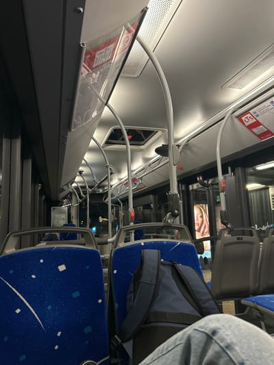 The interior of a bus or train is depicted with rows of blue seats featuring a speckled pattern. Overhead handrails arch across the aisle. Windows reflect the dark exterior. Advertising screens are visible above the seats. One passenger is partially visible, wearing jeans and with a backpack on the seat. The lighting is bright.
