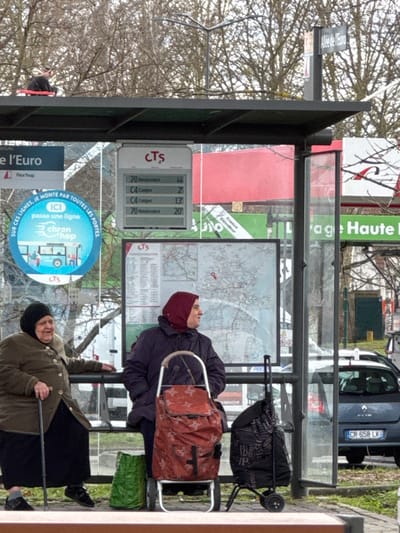 At a bus stop on Allée de l'Euro in Oberhausbergen, France, two women are seated within a transparent bus shelter. The woman on the left, an elderly individual with a black head covering, wears a brown jacket and black skirt, holding a black cane while looking to her left. The woman on the right, wearing a dark purple jacket and a red head covering, is seated in profile, looking towards the right. Next to her are two shopping trolleys: one with an orange and star-patterned bag, and another with a dark bag. A green bag rests on the ground beside the orange trolley.
Above the women, an electronic display board shows "CTS" and bus route information (lines 70 and C4), with a bus network map below it. A blue circular sign promoting "Chron'hop" is visible to the left. A separate sign to the right confirms the location as "Allée de l'Euro".
Above the bus shelter's dark roof, an individual is seated, wearing dark clothing and appearing to have a red object beside them. The background features bare trees, a dark blue Renault Clio car (license plate CR-658-LK), and signage for what appears to be a service station or car wash facility ("Auto," "Lavage Haute Pression").