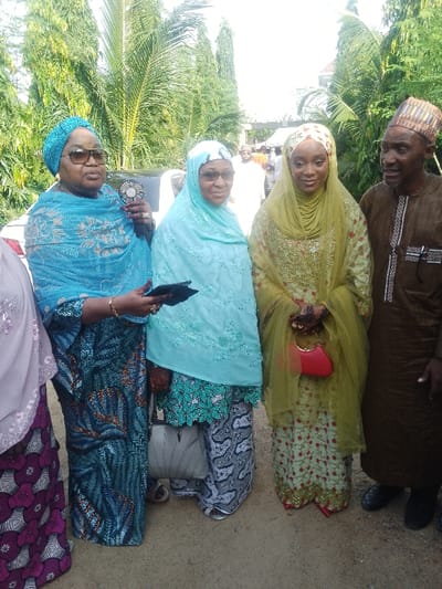 This image captures five individuals—four women and one man—standing outdoors in Unguwa Uku Kauyen Alu, Nigeria, during daylight. From the left, a partial view of a woman is visible, wearing a light purple head covering and a patterned purple and white garment. Next, a middle-aged woman wears a sparkling turquoise blue head wrap, dark sunglasses, a patterned blue dress, and holds a black handheld fan in her right hand and a dark, rectangular object in her left. Gold-colored jewelry and henna patterns are visible on her hands. Adjacent to her, another middle-aged woman wears a light aqua hijab, clear glasses, a matching embroidered top, patterned black and white trousers, and carries a light grey patterned handbag. Her right hand also displays henna. To her right, a younger woman wears a lime green hijab and a long, embroidered lime green dress with red floral patterns. Her hands are extensively adorned with henna, and she holds a small, red, round handbag with a gold-colored handle. On the far right, an elderly man is positioned, wearing a brown traditional kaftan with black and white embroidery and a patterned cap (kufi), looking towards the young woman. The ground is unpaved, light brown dirt. The background consists of dense green foliage, including tall palm trees, with a white vehicle partially visible. Distant, blurred figures in white attire and a white canopy tent are also present.