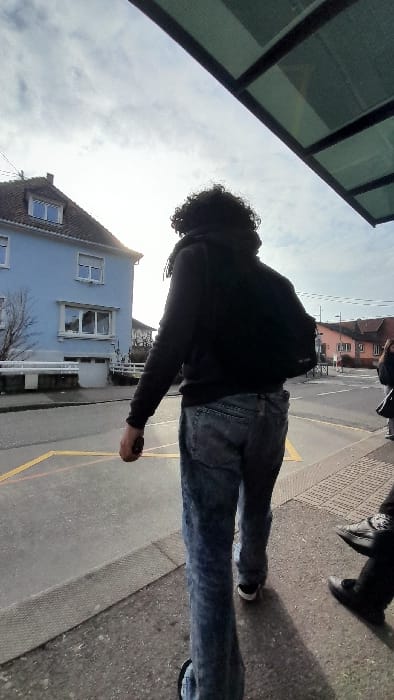 This image depicts an outdoor urban scene at a bus stop in Souffelweyersheim, France, under a bright, partly cloudy daytime sky.
In the foreground, a person is observed from behind, possessing dark, curly hair and wearing a black hooded sweatshirt, faded blue jeans, and black sneakers with white soles. A black backpack is worn on their left shoulder, and a small, dark object is held in their right hand. This individual is positioned on a paved sidewalk, facing away from the camera and looking towards the left side of the frame. To their immediate right and partially out of view, the lower legs and feet of a second individual are visible, clad in dark trousers and black, low-heeled shoes; this person appears stationary.
The upper right portion of the frame shows the green-tinted, translucent roof structure of a bus stop shelter. To the left, a two-story house with light blue exterior walls, white window frames, and a dark tiled roof is prominent, surrounded by bare deciduous trees. A street with yellow geometric road markings separates the sidewalk from the building. Additional residential structures are visible in the distant background on the opposite side of the street.