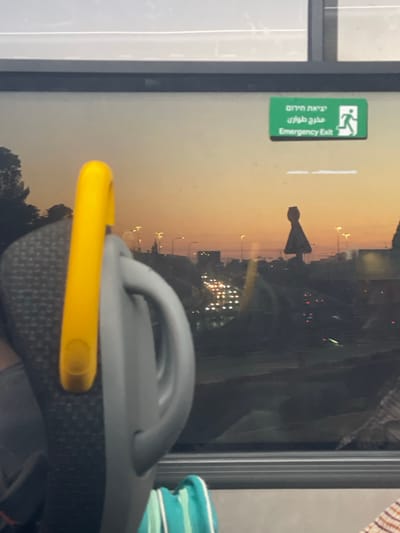A bus window features a reflection of an urban landscape at sunset, including a distinctive monument resembling an inverted cone. A green emergency exit sign is affixed near the window's edge, displaying text in English and Hebrew/Arabic. A portion of a seat with a yellow grab handle is visible in the foreground. Based on the location inferred, the monument visible in the reflection is the Kfar Saba Azrieli Tower.