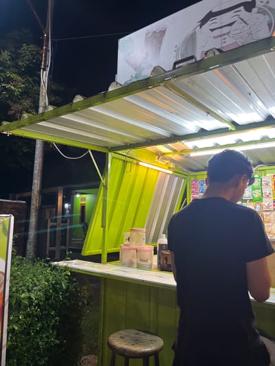 The image depicts an outdoor stall at night in Boja, Indonesia. A person wearing a black t-shirt stands with their back to the camera, facing a stall that sells various packaged goods. The stall has a green metal frame and a corrugated roof illuminated by fluorescent lights. Shelves behind the person are stocked with colorful packages of food items. On the counter, there are several transparent containers with pink lids, a white bottle, and a small wooden dispenser. A stool is visible in the foreground. Lush green bushes are located to the left of the stall, and a building with a green facade and a prominent light fixture is visible in the background. The overall scene suggests a nocturnal commercial setting.